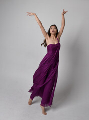 Full length  portrait of pretty brunette asian girl wearing purple flowing  gown. Standing, dancing pose on on studio background.