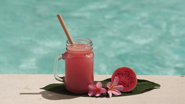 Video footage of glass mason jar with pink guava juice, bamboo straw, half of fresh pink guava, tropical flower frangipani and bubbling blue swimming pool on background.