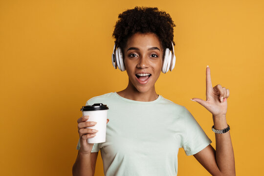 Young black woman with headphones pointing finger and holding coffee