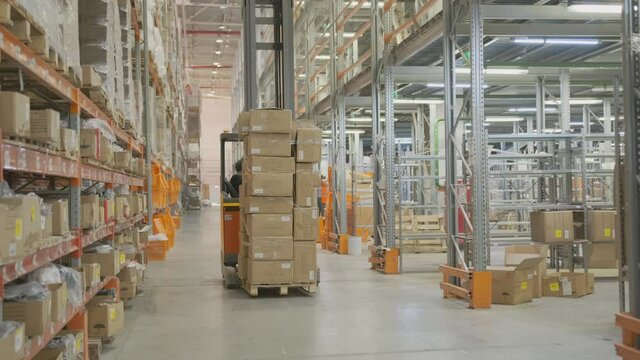 Worker in warehouse rides forklift moving pallets of cardboard boxes of goods between row of racks.