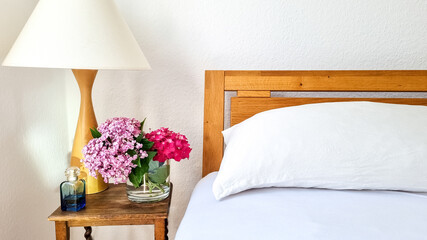 Contemporary natural bedroom with white  silk pillow bedding, a bed next to a side table with pink and purple hortensia flower bouquet in a glass vase, perfume bottle and a white and wooden lamp. 