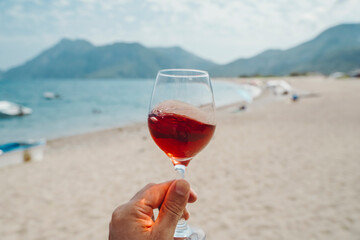 Glass of wine in hand. A glass of red wine against the background of the Mediterranean beach and the sea in a tourist town in the summer under sunlight. Summer, travel, lifestyle, relaxation, and