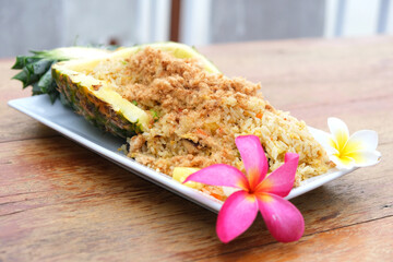 Thai food, pineapple fried rice with shredded pork is on the wooden table with yellow and white frangipani flowers.