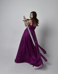 Full length  portrait of pretty brunette asian girl wearing purple flowing  gown. Sitting pose holding a sword  on on studio background.