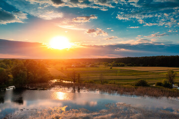 Beautiful sunset in the countryside in summer with the blazing sky. Rural landscape in the evening. Aerial view of brook, village, and arable fields. Panoramic view