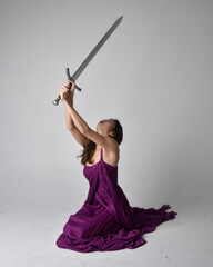 Full length  portrait of pretty brunette asian girl wearing purple flowing  gown. Sitting pose holding a sword  on on studio background.