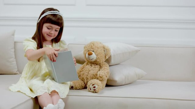 girl with teddy bear reading a book while sitting on the couch