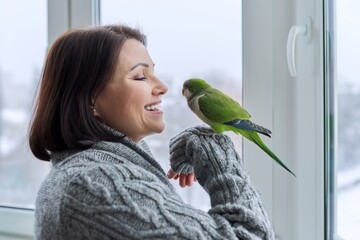 Middle aged woman and parrot together, female bird owner talking looking at green quaker pet - obrazy, fototapety, plakaty