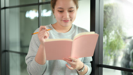 A teenage asian woman with a pen stands by the window and looks down at the notebook in her hand.