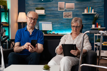 Senior married couple using smartphone and tablet devices with online internet modern technology in living room on sofa. Retired caucasian mature family with walk frame and digital devices