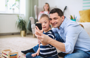 Father with happy down syndrome son indoors at home, taking selfie with smartphone.