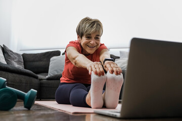Senior woman doing online yoga workout class on laptop computer at home - Sport, technology and healthy elderly lifestyle concept - Focus on face