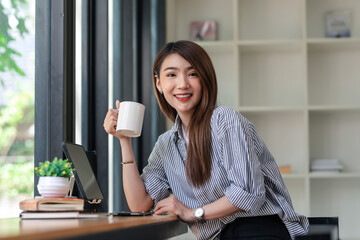 Charming young Asian businesswoman sitting at work drinking coffee. Looking at camera.