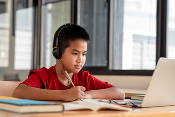 Image of a male student studying online wearing headphones and taking notes on his laptop placed in his room.