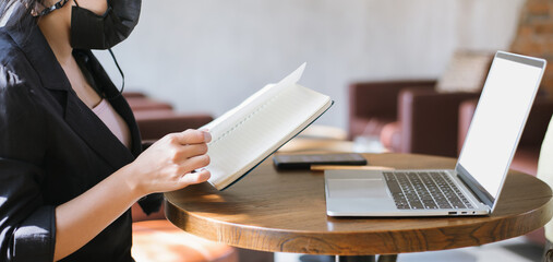 Young asian businesswoman wearing face mask holding book and using laptop work in cafe