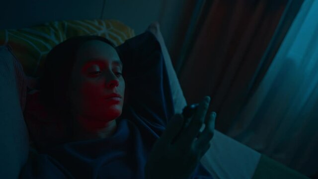 Young beautiful woman lying on bed alone in dark bedroom with hand under her arm. She takes her smartphone to check it up and looks at screen. Modern technology concept. Slow motion cinematic shot