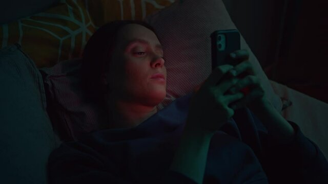 Beautiful girl typing on her smartphone while lying on bed alone in dark bedroom. Young woman using mobile device for surfing and chatting. Modern technology concept. Slow motion cinematic shot