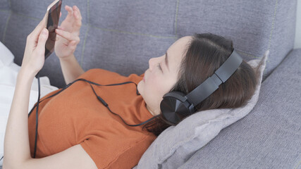 attractive modern asian woman listening music in headset on sofa in living room at home. portrait happy female relaxing playing song playlist from smartphone via headphone. entertainment technology.