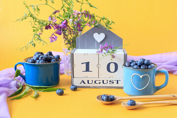 Calendar for August 10 :the name of the month of August in English, cubes with the number 10, blueberries in blue cups, bouquets of flowers, yellow background, side view