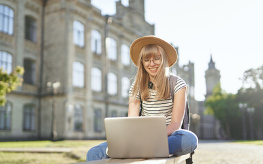 Young happy blonde school girl, college or university student with backpack and hat using laptop sitting on bench in university campus. Browsing Internet, chatting, education or surfing concept