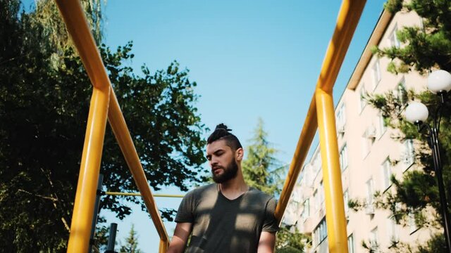 Athletic male is resting on ground near horizontal bar. Caucasian man with beard and dreadlocks stands outside next to parallel bars and takes breath before starting strength training.