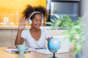 Little girl studying online using her laptop at home. Beautiful young girl with headset is sitting in front of her laptop during corona time. Learning remotely during covid coronavirus pandemic