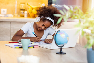 Portrait of smiling little biracial girl in headphones do homework study online in kitchen, happy small African American child in earphones have online web class or lesson using laptop at home