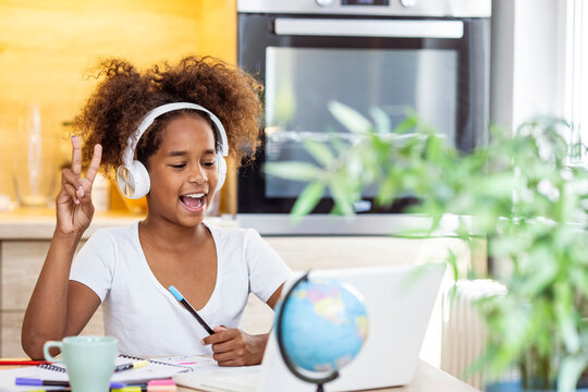 Smiling small African American girl in headphones watch video lesson on computer in kitchen, happy little biracial child in earphones have online web class using laptop at home, homeschooling concept