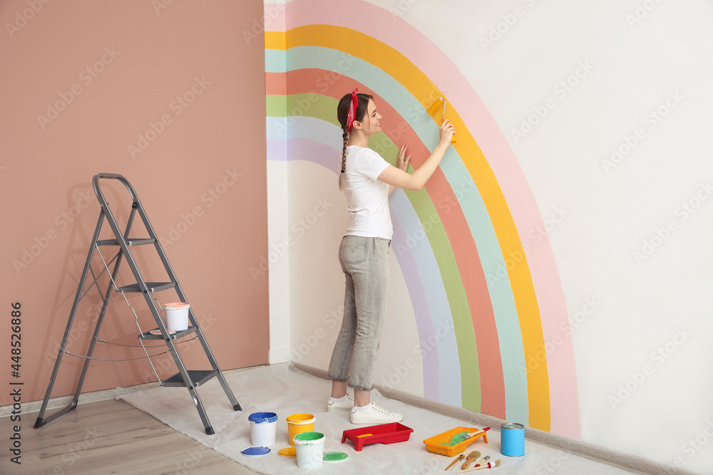 Wall mural Young woman painting rainbow on white wall indoors