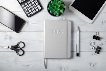 Minimal  unisex workplace with tablet,  silver notepad diary 2022, smartphone, small succulent and stationery on a light wooden background.