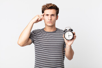 young handsome man feeling confused and puzzled, showing you are insane and holding an alarm clock