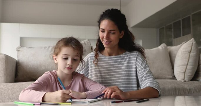Adorable interested little child daughter drawing in paper album with caring millennial mother or nanny, spending weekend free time together at home, domestic leisure hobby activities concept.