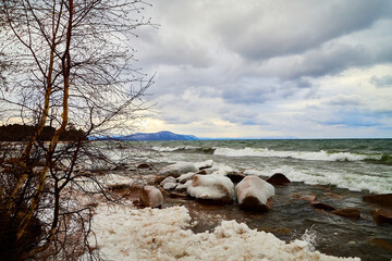 Nature landscape with dramatic sky and clouds, lake with ice, shore with snow and dark trees in...