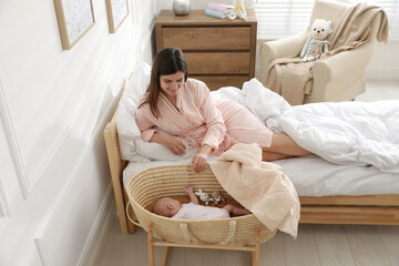Happy young mother resting near cradle with sleeping newborn baby at home
