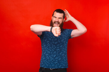 Angry and unpleasant face of bearded man is looking at his watch and rising hand over red background