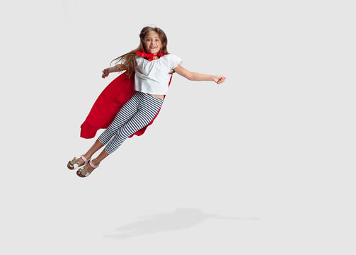 Full-length portrait of beautiful little girl flying like super hero in red cape isolated on gray studio background. Childhood, education, emotion concept. Flyer