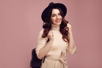 Charming lady with a cute look dressed in stylish casual clothes, wearing a black hat and backpack, smiling sincerely against pink isolated