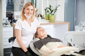 Horizontal snapshot of pretty, young female dentist, wearing uniform, rubber gloves, smiling, sitting at her working place in well equipped dental office. Female patient during dentist's visit