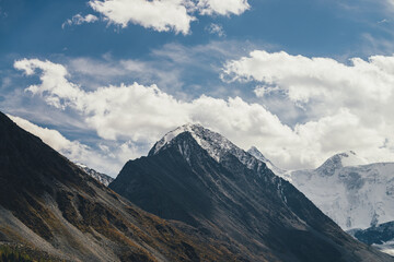 Atmospheric alpine landscape with high mountain silhouette with snow on peaked top under blue cloudy sky. Dramatic mountain scenery with beautiful snow-covered pointy peak and high snowy mountain wall