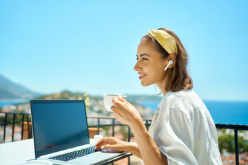 Young business woman drinking coffee and looking away, working on laptop computer at morning while sitting at terrace with sea view on resort
