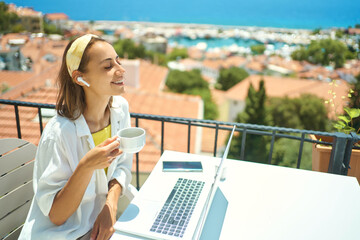 Fototapeta na wymiar Pleased woman enjoying morning coffee while sitting on terrace with sea view on Turkish resort, using laptop computer for remote work, freelance concept