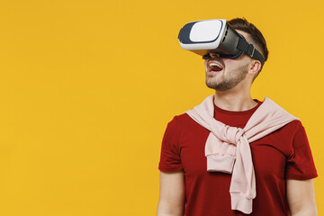 Excited impressed amazed happy young man 20s wear red t-shirt casual clothes watching in vr headset pc gadget isolated on plain yellow color wall background studio portrait. People lifestyle concept.