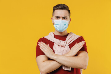 Young man in red t-shirt sterile face mask ppe to safe from coronavirus virus covid-19 flu on lockdown quarantine do stop crossed hand gesture isolated on plain yellow wall background studio portrait