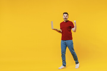 Full length fun smiling young man in red t-shirt casual clothes using laptop pc computer work clench fist do winner gesture celebrate isolated on plain yellow color wall background studio portrait