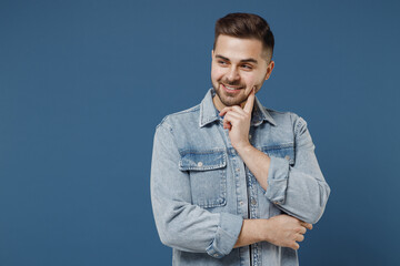 Charming young handsome brunet man 20s wears denim jacket put hand prop up on cheek lost in thought and conjectures isolated on dark blue background studio portrait. People emotions lifestyle concept.