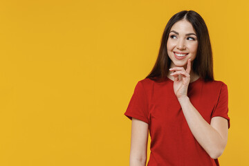 Smiling young brunette woman 20s wears basic red t-shirt put hand prop up on chin lost in thought and conjectures look up above isolated on yellow background studio portrait. People emotions concept.