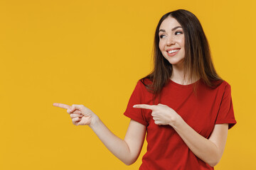 Smiling joyful young brunette woman 20s wears basic red t-shirt point look aside on workspace area copy space mock up isolated on yellow background studio portrait. People emotions lifestyle concept.