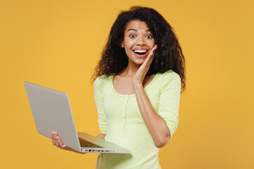 Shocked surprised stupefied amazed african american young brunette woman 20s wear green shirt hold use work on laptop pc computer keeping mouth wide open isolated on yellow background studio portrait