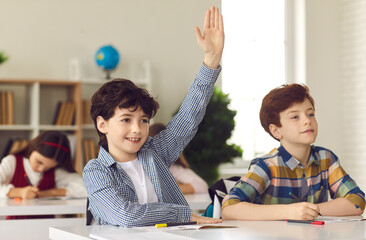 Smart schoolboy sits at a desk next to his classmate in class with his hand raised, wanting to give...