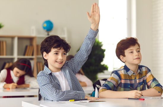 Smart schoolboy sits at a desk next to his classmate in class with his hand raised, wanting to give the right answer or participate. Education, primary school, learning and people concept.
